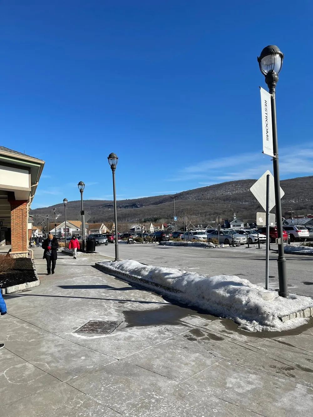 The image shows a sunny day with clear blue skies over a paved area with some snow on the ground street lamps and a sign pointing to restrooms with hills in the distance and people walking nearby