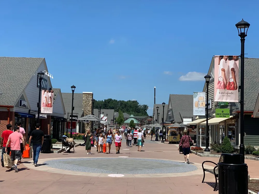 Shoppers walk through an outdoor shopping village with various brand stores on a sunny day