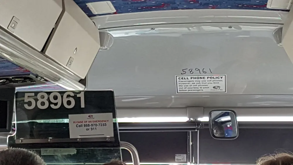 The image shows the interior front area of a bus with emergency contact information and a cell phone policy notice displayed above the windshield