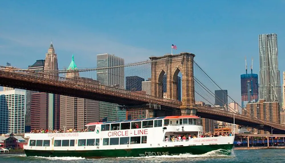A Circle Line sightseeing cruise boat is filled with passengers as it passes under the Brooklyn Bridge with the New York City skyline in the background