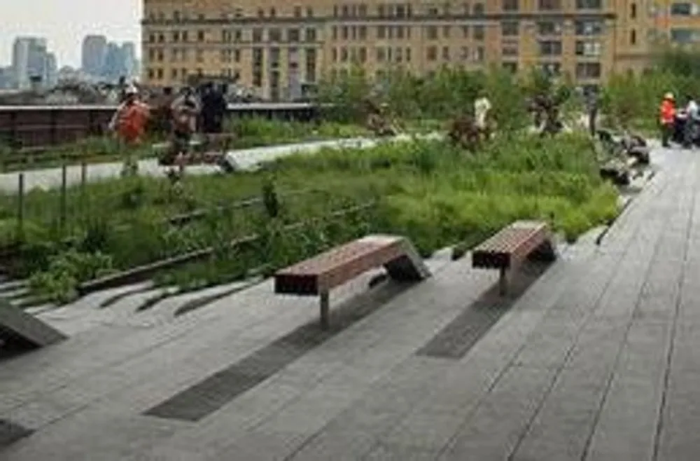 The image shows a modern urban park with wooden benches and lush greenery against a backdrop of city buildings with people walking along the path