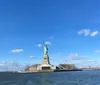 The photo captures the Statue of Liberty standing on Liberty Island against a backdrop of clear blue sky with scattered clouds viewed from the perspective of the surrounding water
