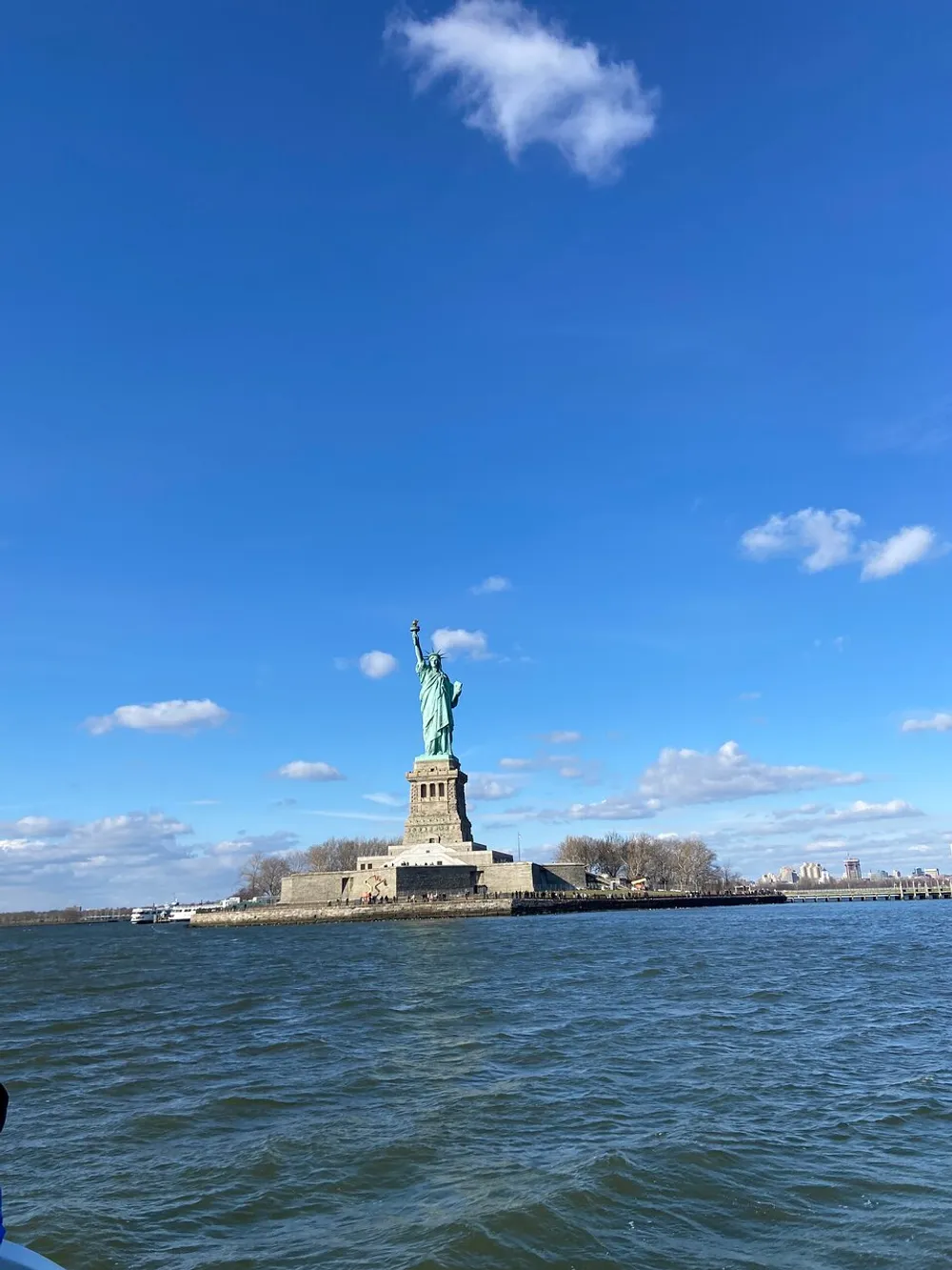The photo captures the Statue of Liberty standing on Liberty Island against a backdrop of clear blue sky with scattered clouds viewed from the perspective of the surrounding water