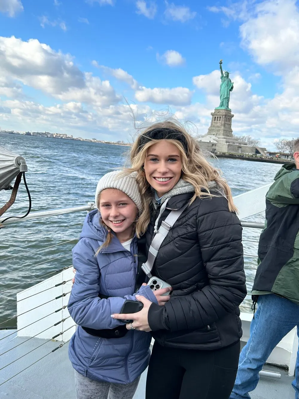 Two people are smiling for a photo on a boat with the Statue of Liberty in the background