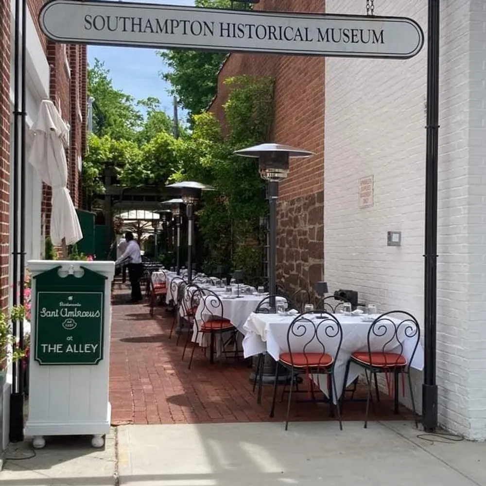 The image shows an outdoor dining setup with white cloth-covered tables and red chairs along a narrow alley beside the Southampton Historical Museum as indicated by the overhead sign with a waiter in the background and outdoor heaters present