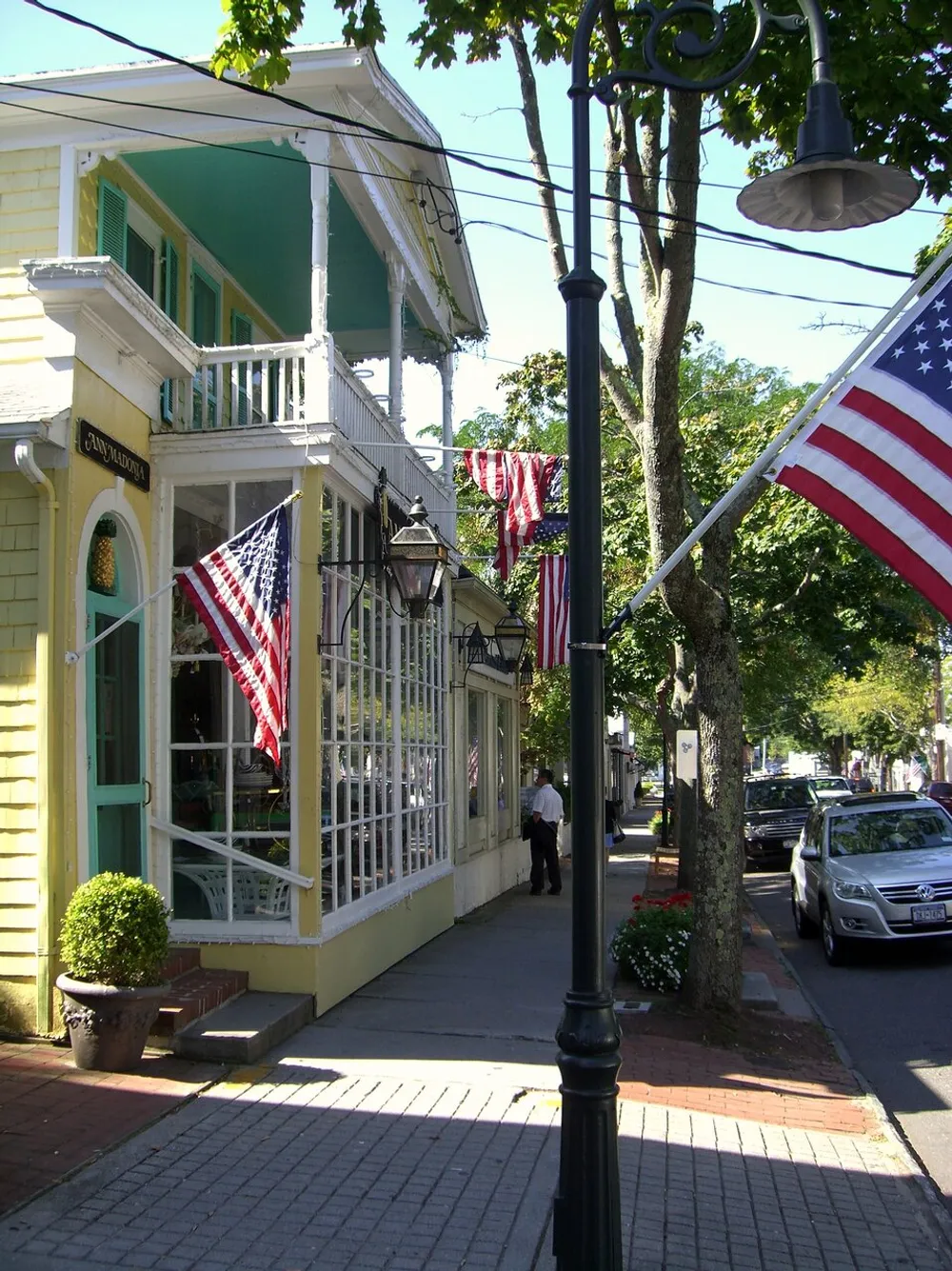 An American flag-adorned street scene featuring a person walking by a quaint yellow building with a green balcony street lamps and parked cars