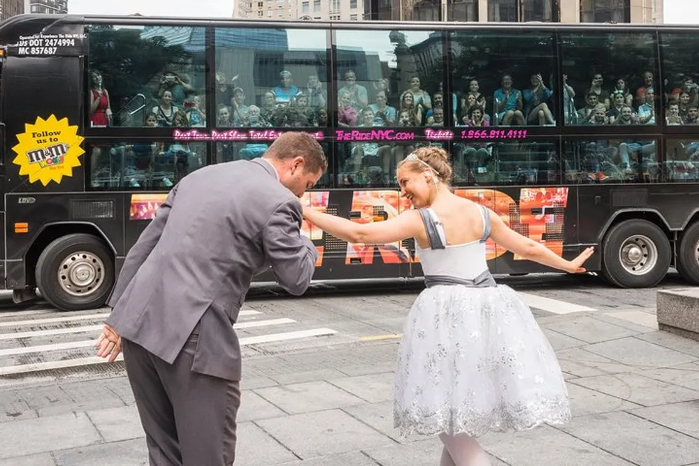 A man bows to a joyful woman in a dress as amazed passengers on a tour bus look on
