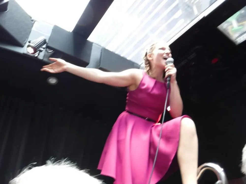 A person in a pink dress is photographed from a low angle while singing into a microphone and gesturing with one arm outstretched