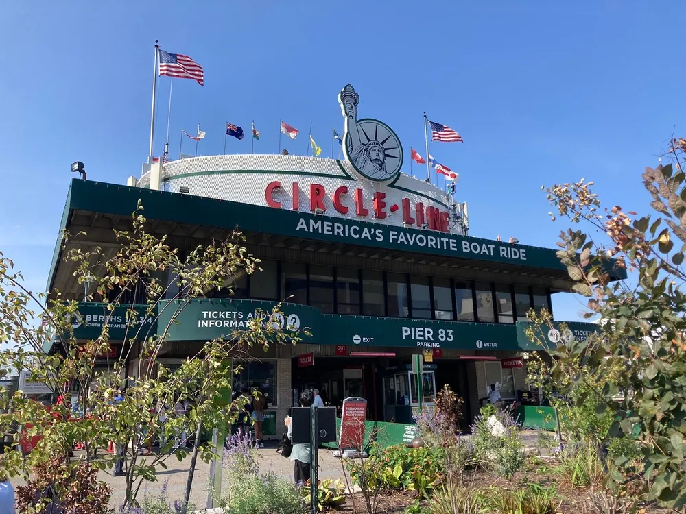 The image shows the Circle Line ticket booth at Pier 83 advertised as Americas Favorite Boat Ride flanked by plants and flying the American flag under a clear blue sky