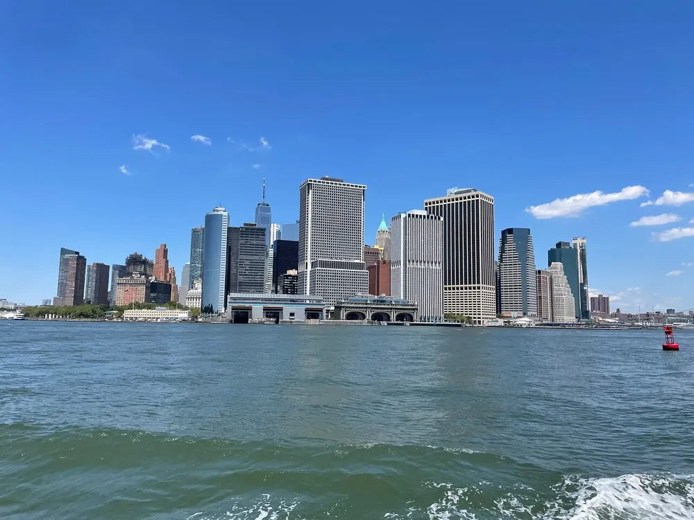 The image shows a clear view of the Lower Manhattan skyline seen from the water under a bright blue sky