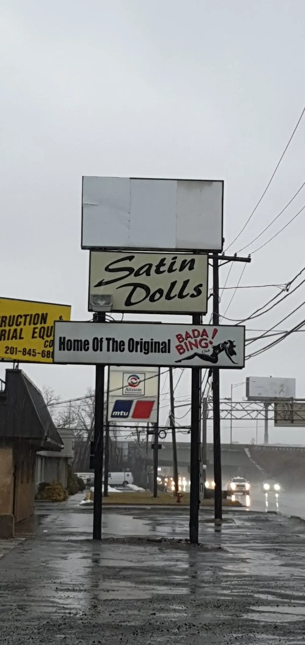 The image shows a vertical roadside sign for Satin Dolls advertised as the Home Of The Original BADA BING on a rainy day with street reflections and headlights from approaching traffic
