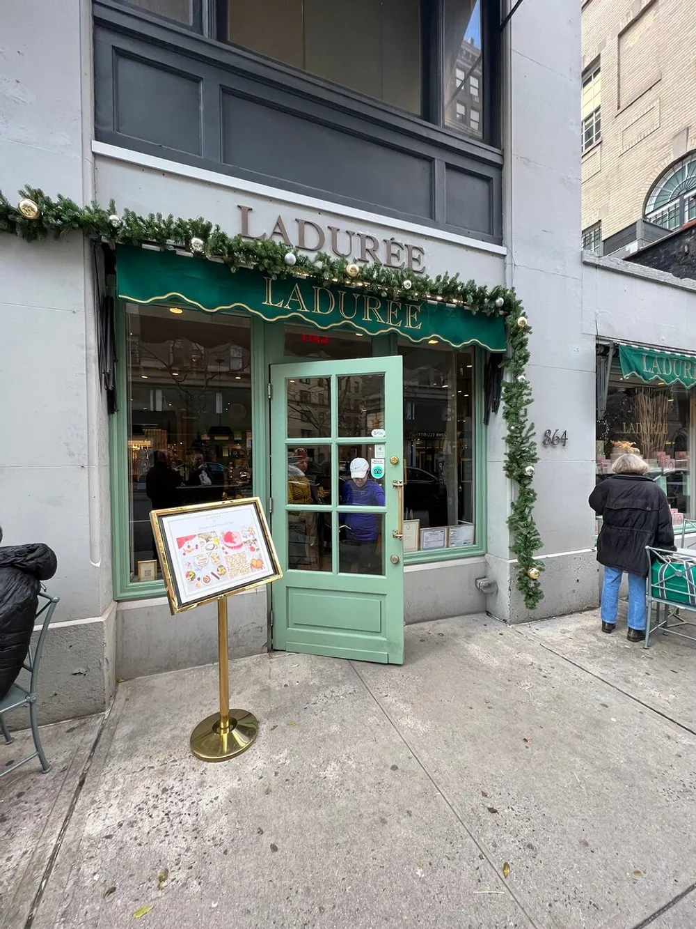 The image shows the exterior of a Ladure patisserie decorated with festive greenery featuring its pastel green storefront with a menu stand outside and a glimpse of the interior where customers are present