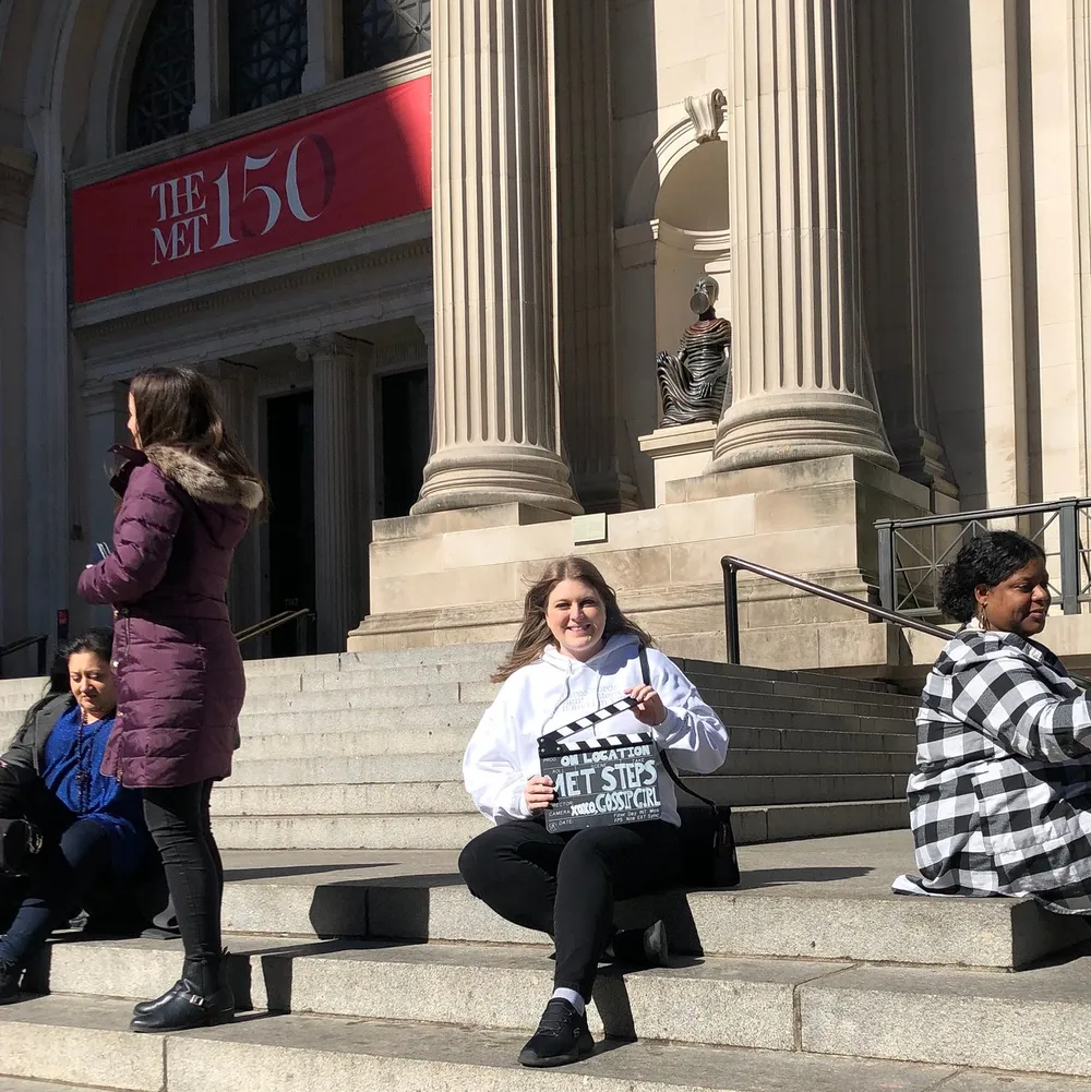 Several people are sitting on the steps of a building with the banner THE MET 150 suggesting it is the entrance to the Metropolitan Museum of Art during its 150th anniversary