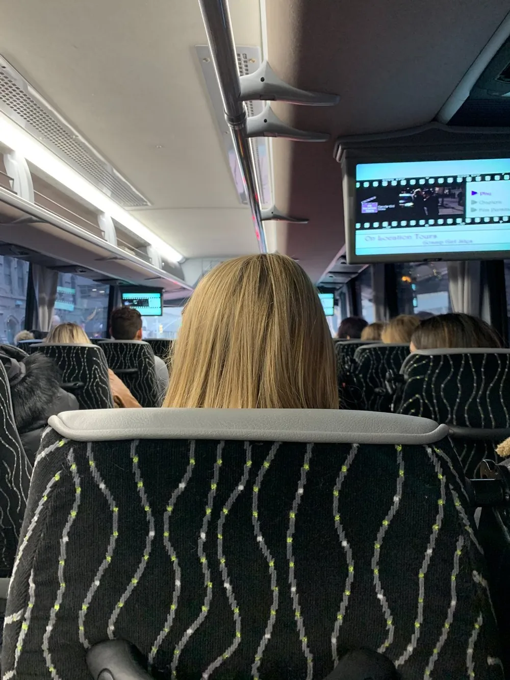 A person with blonde hair is seated in a bus facing forward with a screen displaying some information above them