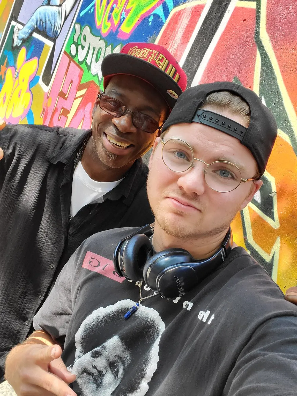 Two smiling men are taking a selfie in front of a colorful graffiti wall