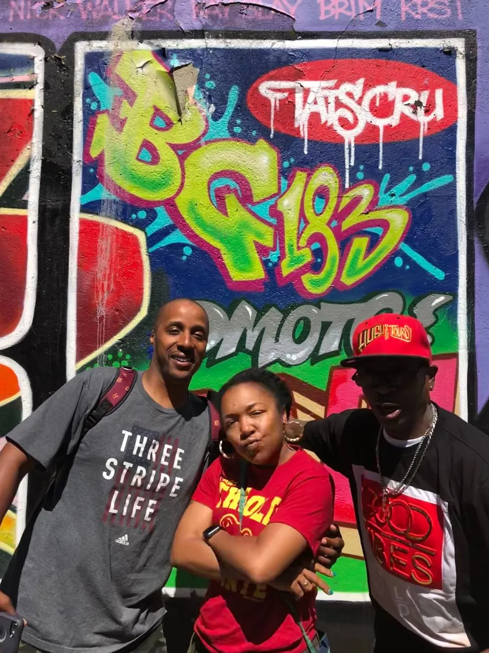 Three people are posing with confident expressions in front of a colorful graffiti wall
