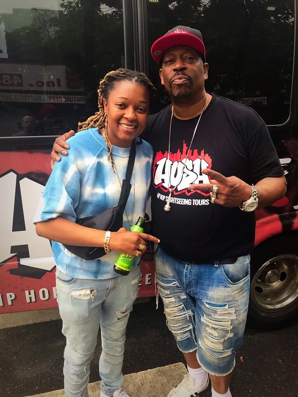 Two individuals are posing for a photo together on the street with a hip hop sightseeing tour bus in the background and one of them holding a drink