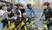 A group of cyclists wearing helmets pauses during a bike tour to listen to their guide, with a city skyline and water as the backdrop.