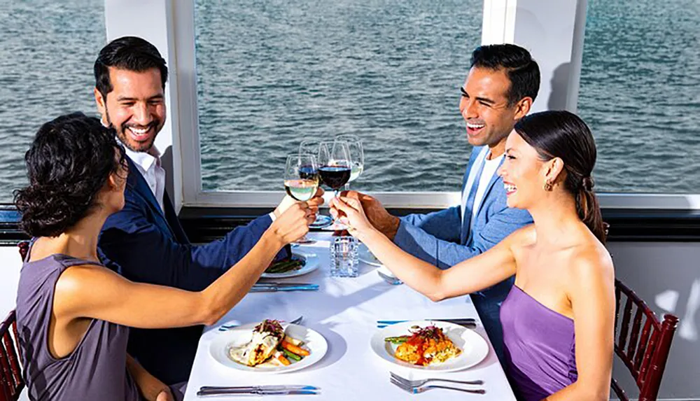 Four people are cheerfully toasting with glasses of wine at a dining table by a window overlooking the water