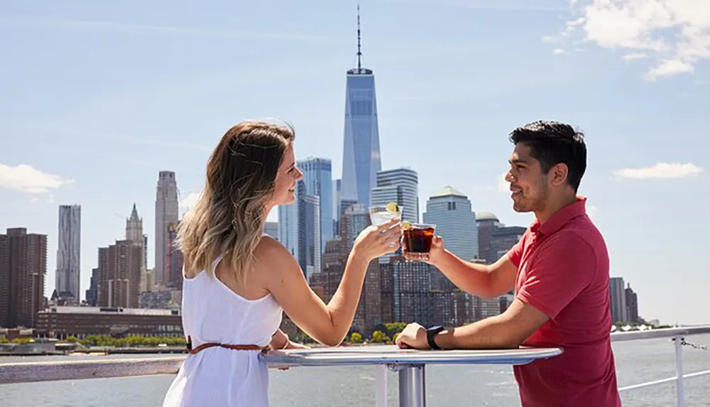 A man and a woman are toasting drinks at a railing with a city skyline in the background