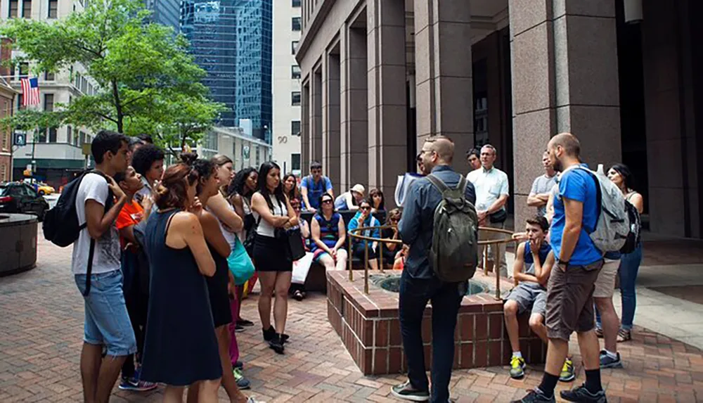 A group of people is attentively listening to a speaker outdoors possibly during a guided tour or outdoor lecture in an urban setting