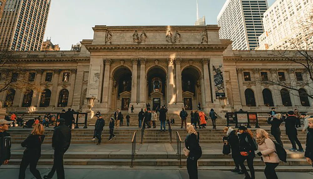 People are seen walking up and down the steps of a grand historic building with classical architecture likely a library or museum bathed in the warm light of a setting or rising sun