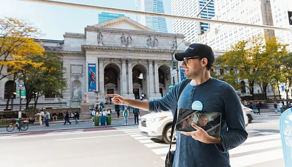A man wearing a cap and holding a tablet is pointing towards something in the direction of a city street with the classical facade of a grand building possibly a museum in the background