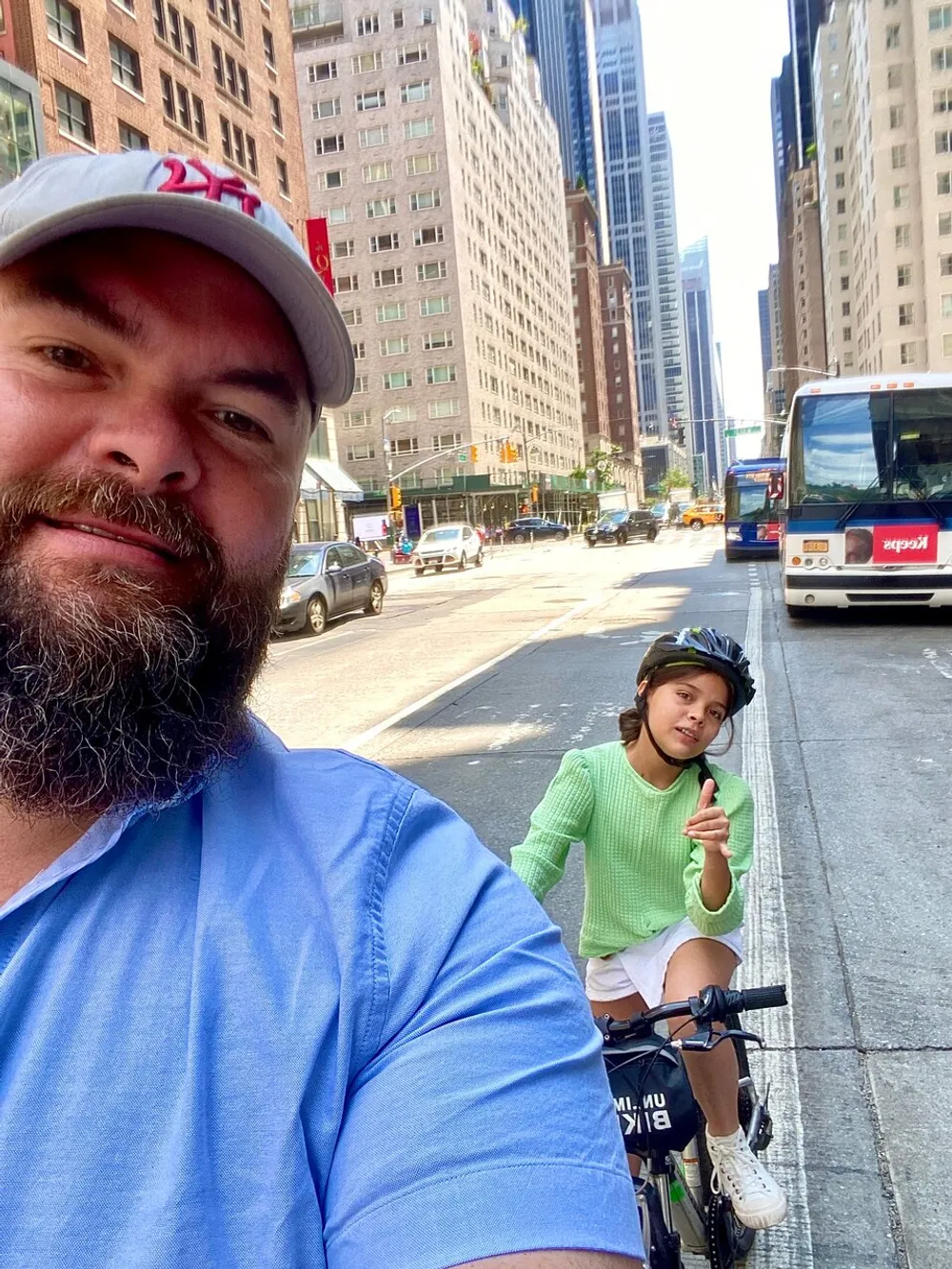 A person takes a selfie while another person rides a bike behind them on a city street