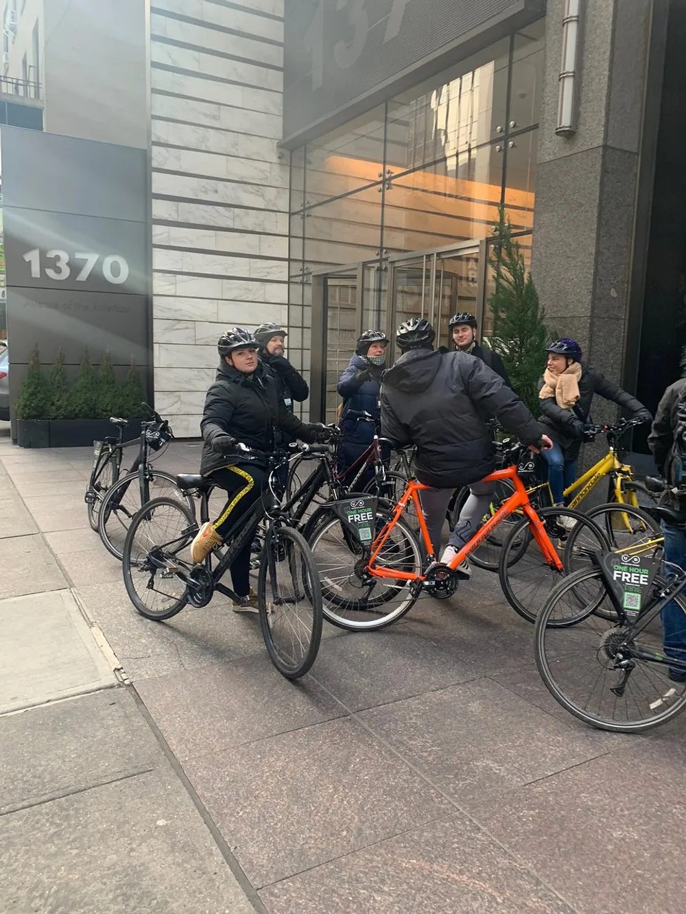 A group of cyclists wearing helmets is gathered on a city sidewalk with some bikes featuring branding suggesting they may be part of a bike-sharing program