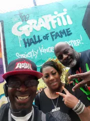 Three people are posing with smiles and hand signs in front of a colorful wall with graffiti that reads 