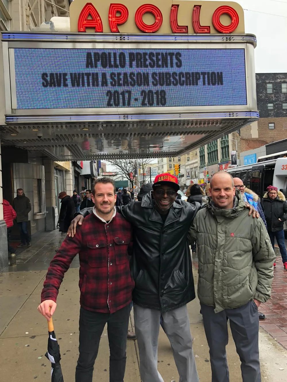 Three smiling men are posing together in front of the iconic Apollo Theater marquee which advertises a season subscription for 2017-2018