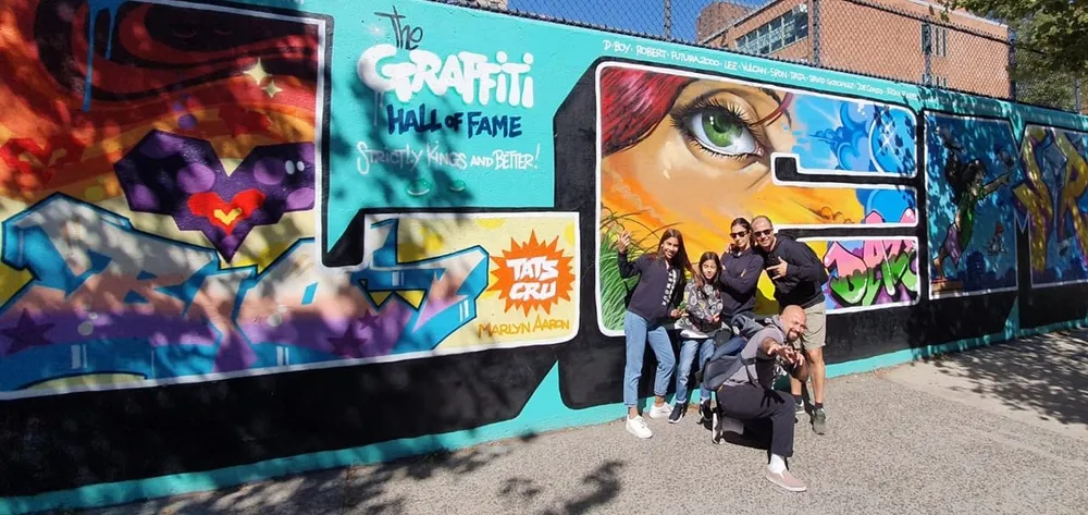 A group of people is posing for a photo in front of a vibrant graffiti wall that includes the text The Graffiti Hall of Fame colorful artwork and a large depiction of an eye