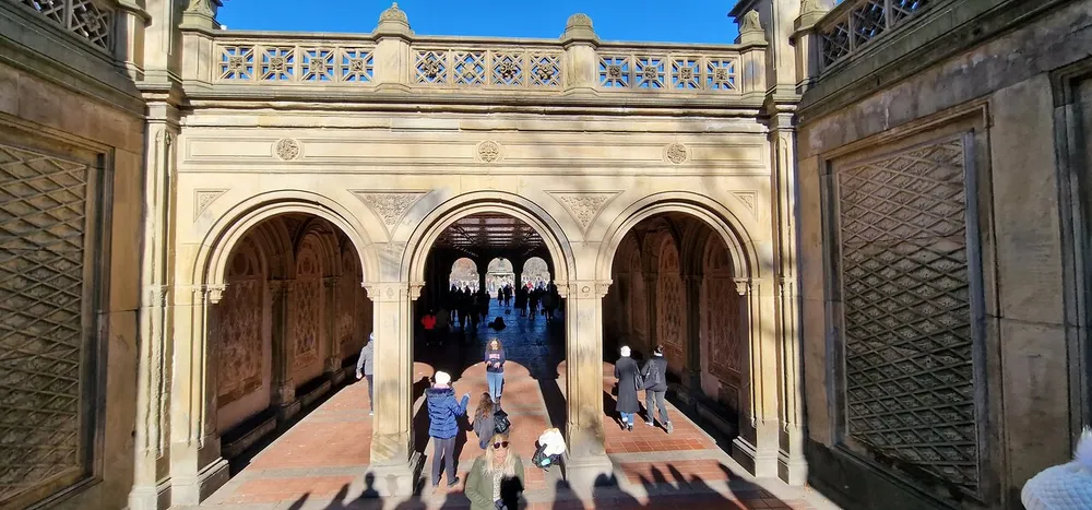 The image shows people walking through an ornate arched walkway with sunlight casting shadows possibly within a historical or park setting