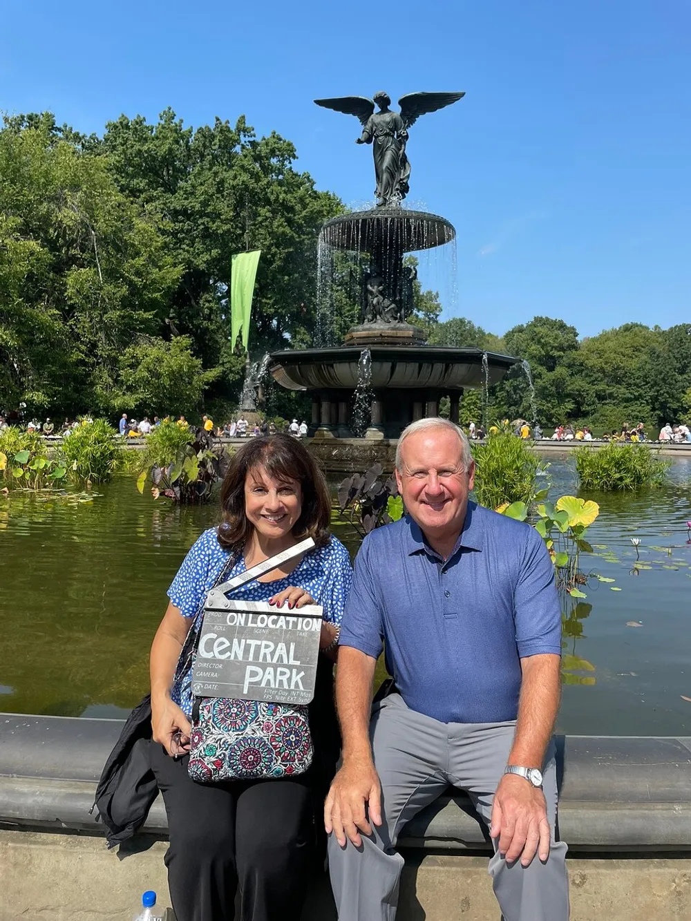 A smiling man and woman pose in front of the Bethesda Fountain in Central Park with the woman holding a sign that reads On Location Central Park