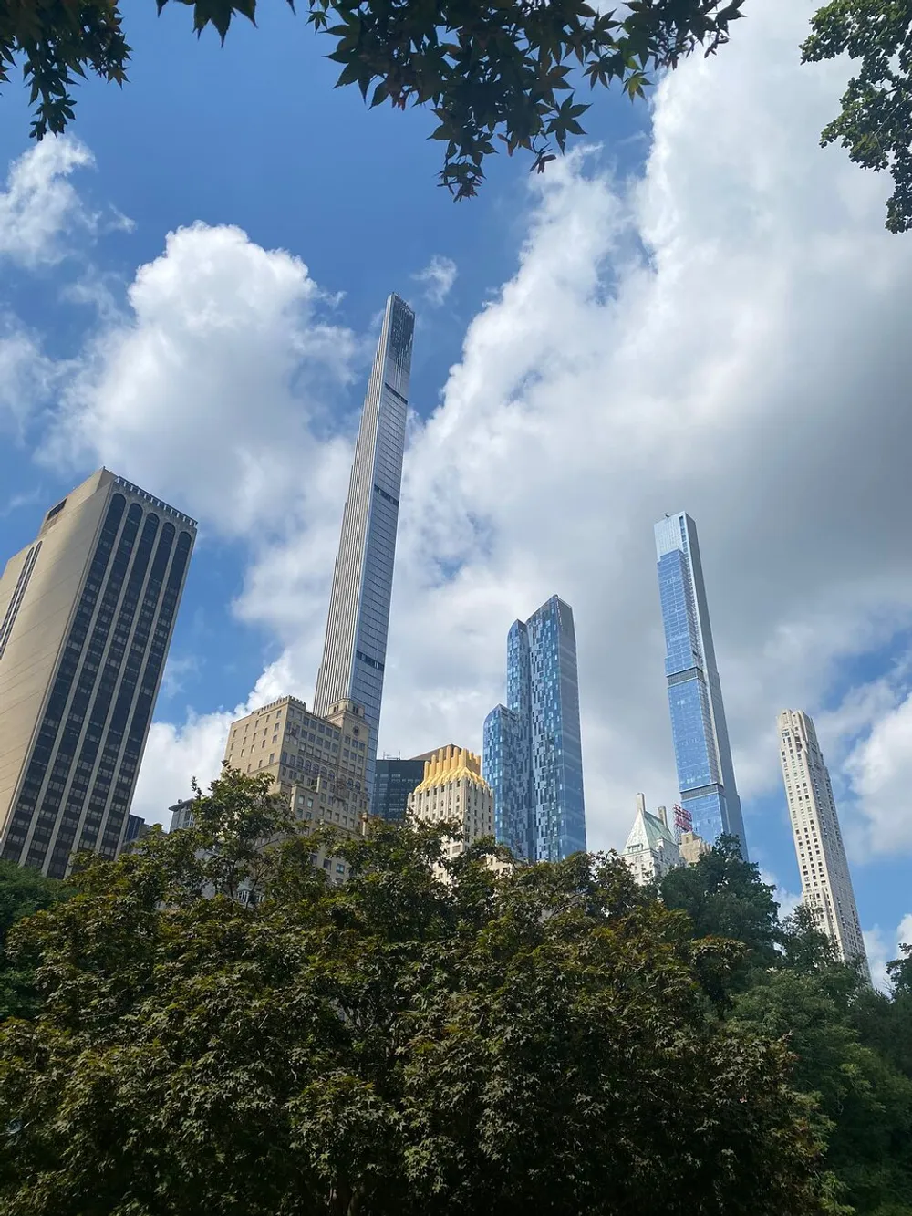 The image captures a view of tall skyscrapers soaring into the sky juxtaposed with the natural greenery of treetops in the foreground under a cloudy blue sky