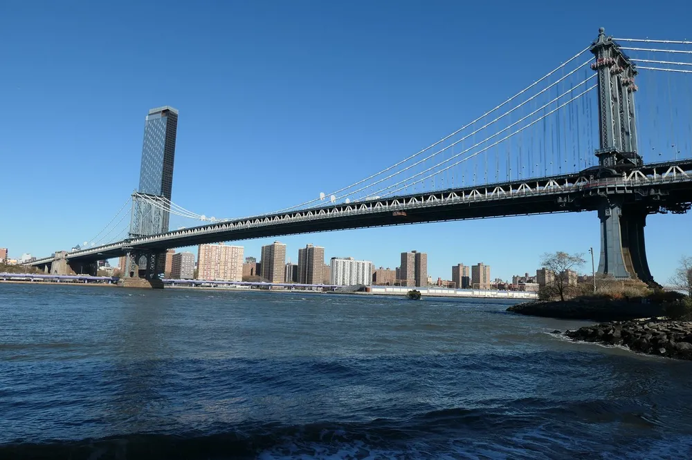 This is a daytime image of the Manhattan Bridge spanning over the East River with clear skies and the skyline of apartment buildings in the background