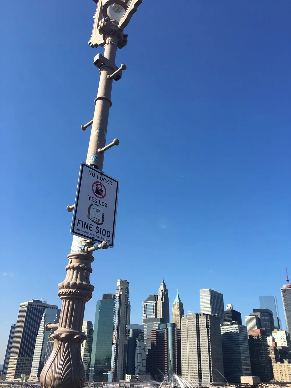 The image shows a lamppost with two conflicting signs about locks one stating NO LOCKS the other YES LOX with a humorous fine warning set against a clear blue sky and a backdrop of a city skyline