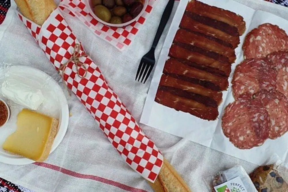 The image shows a selection of picnic foods like slices of cheese sausage a baguette and condiments laid out on a tablecloth with a classic red and white checkered pattern