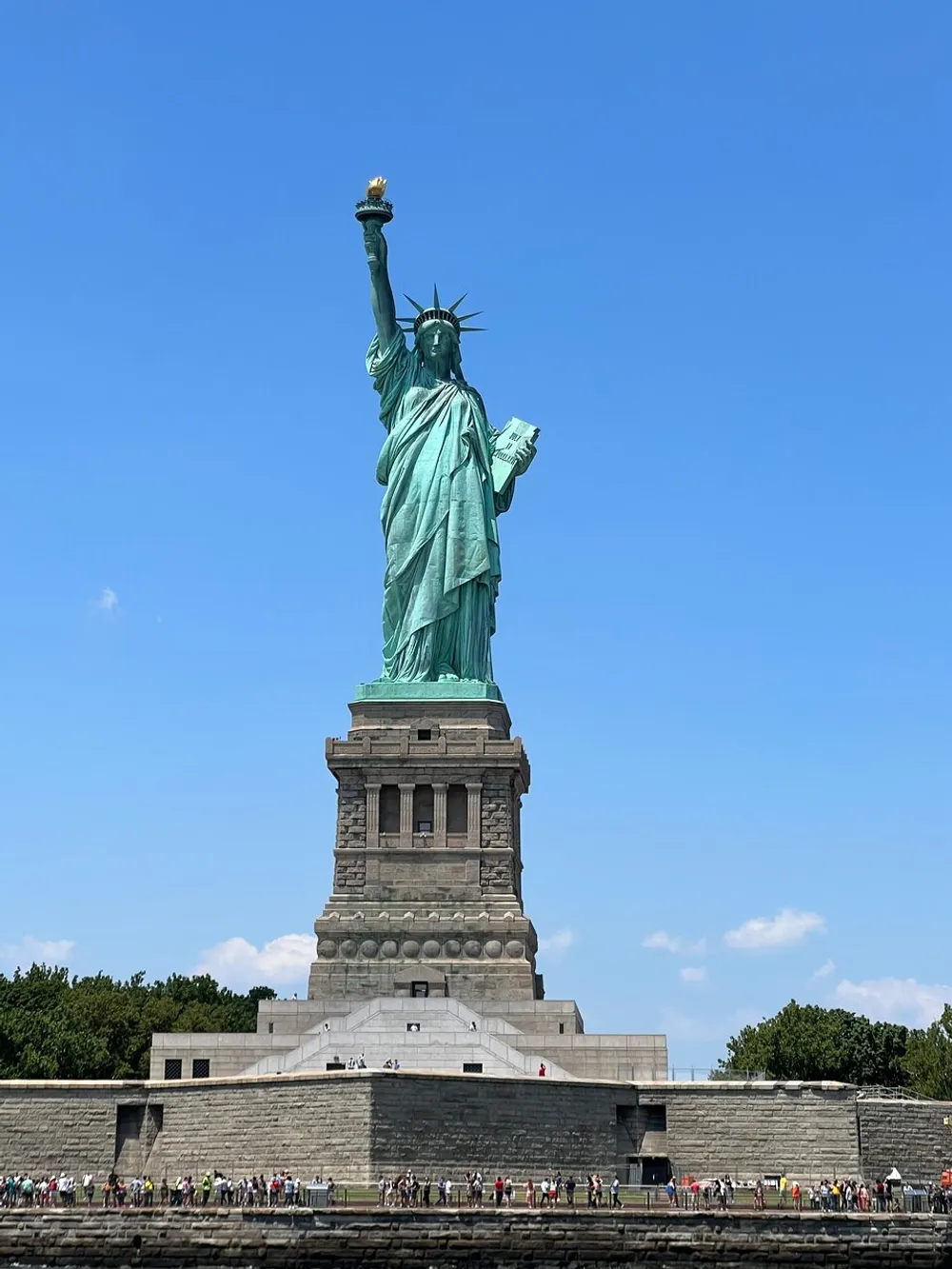 The image shows a full view of the Statue of Liberty against a clear blue sky with visitors visible at its base on Liberty Island