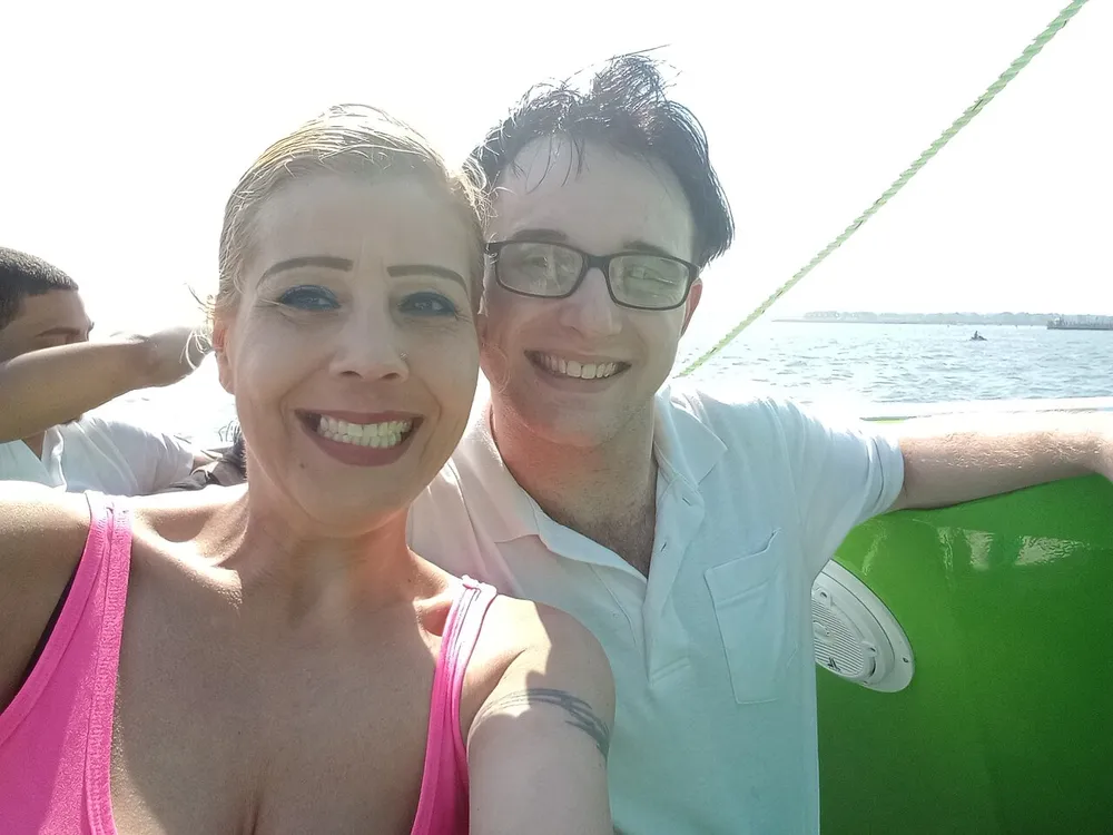 A man and a woman are smiling for a selfie on a sunny day possibly on a boat given the glimpse of water in the background and the presence of a lifebuoy