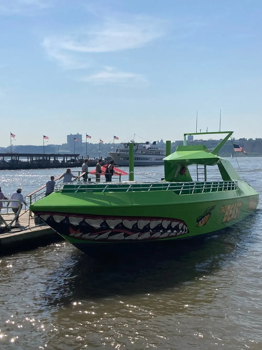 The image shows a green tour boat with shark teeth graphics docked at a pier with passengers aboard on a sunny day