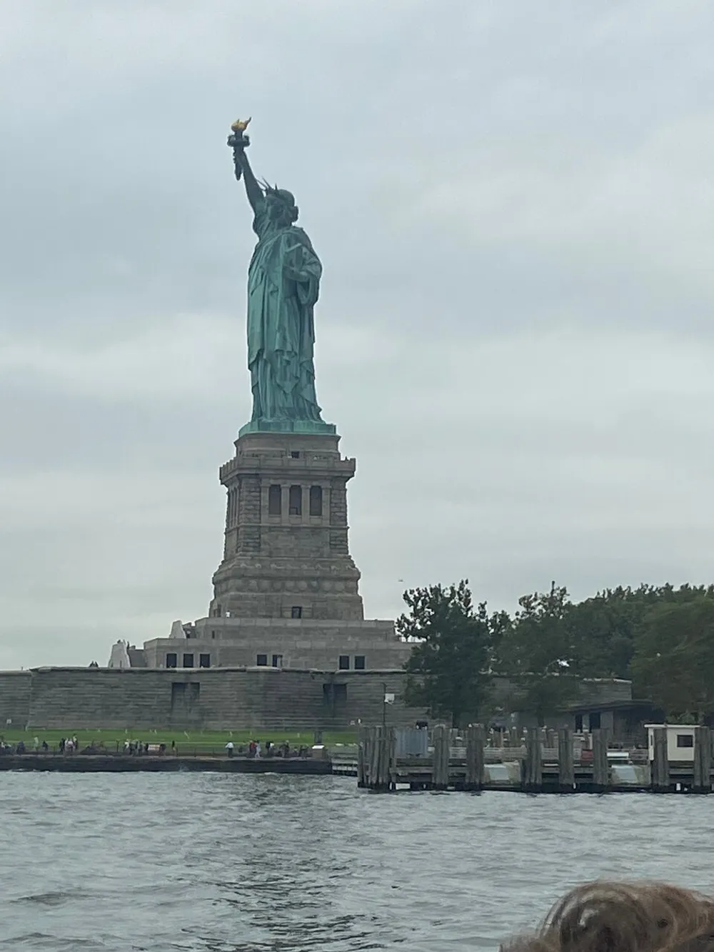The Statue of Liberty is seen standing tall against an overcast sky viewed from the water with a glimpse of visitors near its base
