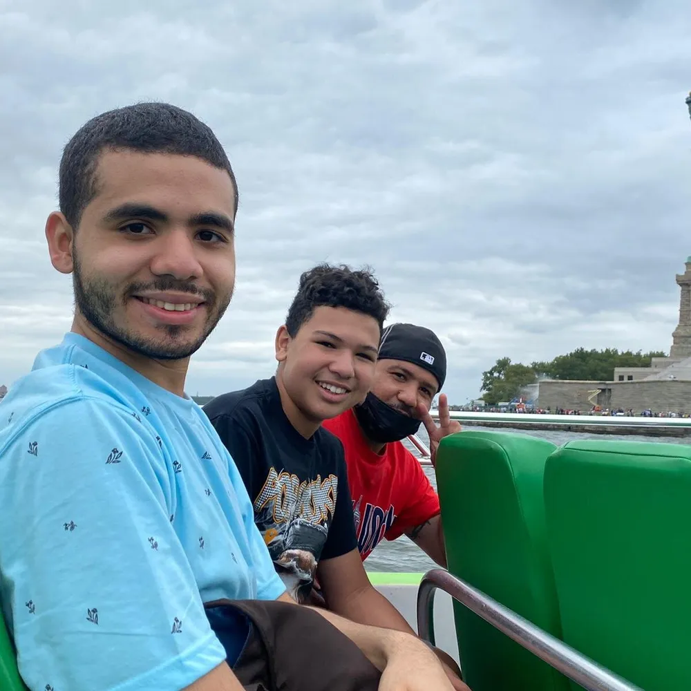 Three people are posing for a photo with smiles aboard a boat with a cloudy sky above and what appears to be a monument in the background