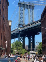This image features pedestrians and vehicles on a bustling street framed by old brick buildings with the iconic steel structure of the Manhattan Bridge towering in the background under a clear blue sky.