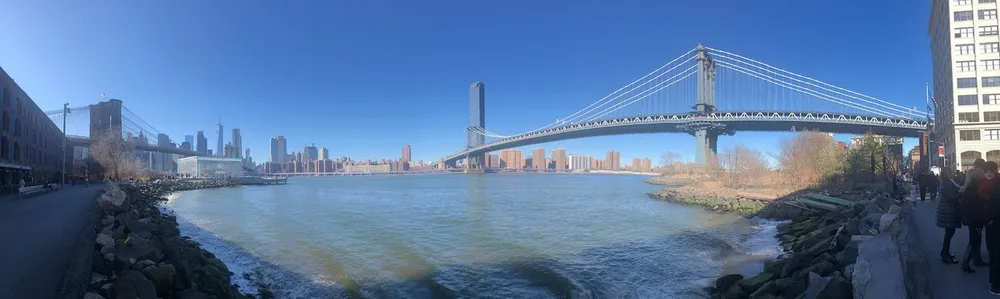 A panoramic view of the Manhattan Bridge spanning over the East River with the New York City skyline in the background and people enjoying the riverside walkway