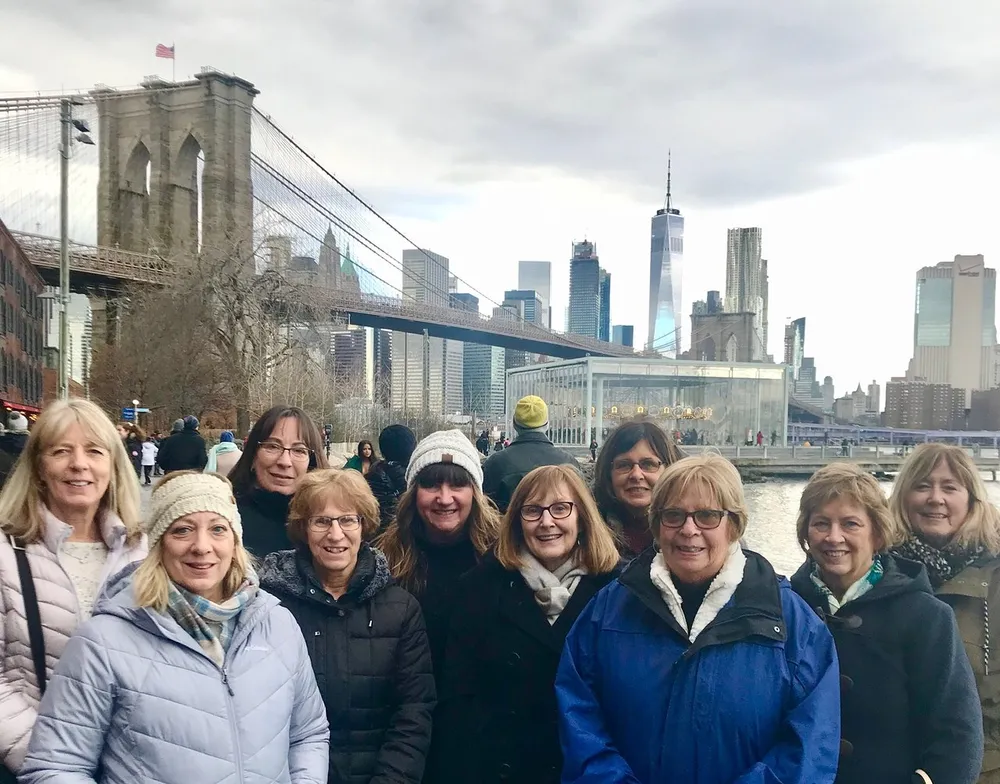A group of smiling people is posing for a photo with the Brooklyn Bridge and the Manhattan skyline in the background