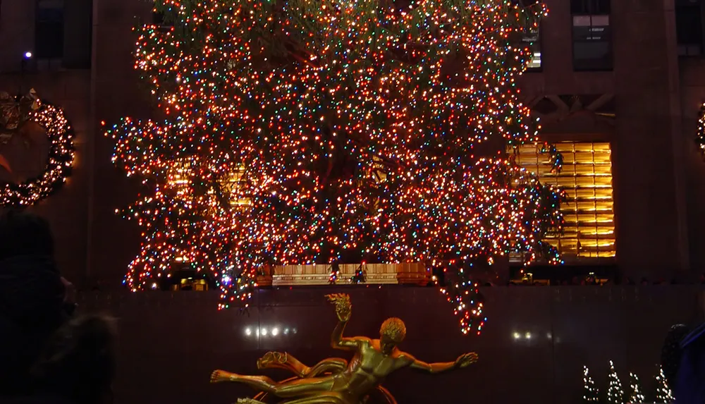 A large brightly lit Christmas tree stands above a golden statue possibly depicting a scene of holiday festivity in an urban setting