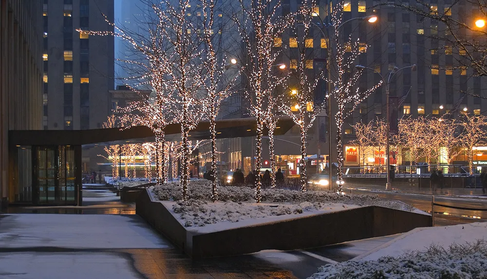 The image shows a winter evening with snow-covered sidewalks and trees adorned with twinkling lights alongside an urban street