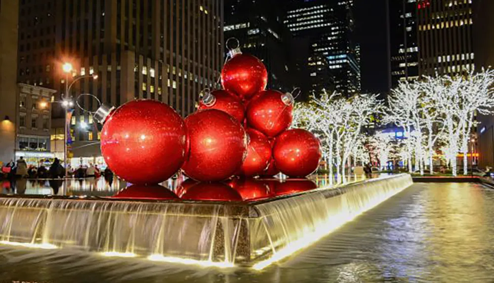 An outdoor holiday display features oversized red Christmas ornaments amidst illuminated trees at night in an urban setting