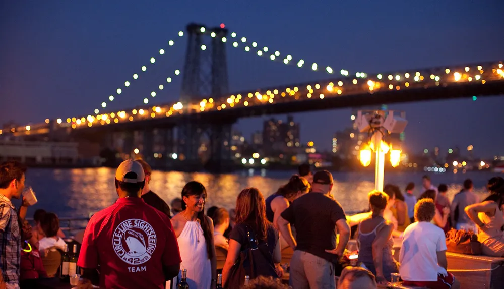 A group of people enjoys an evening by the water with a lit bridge in the background creating a picturesque scene